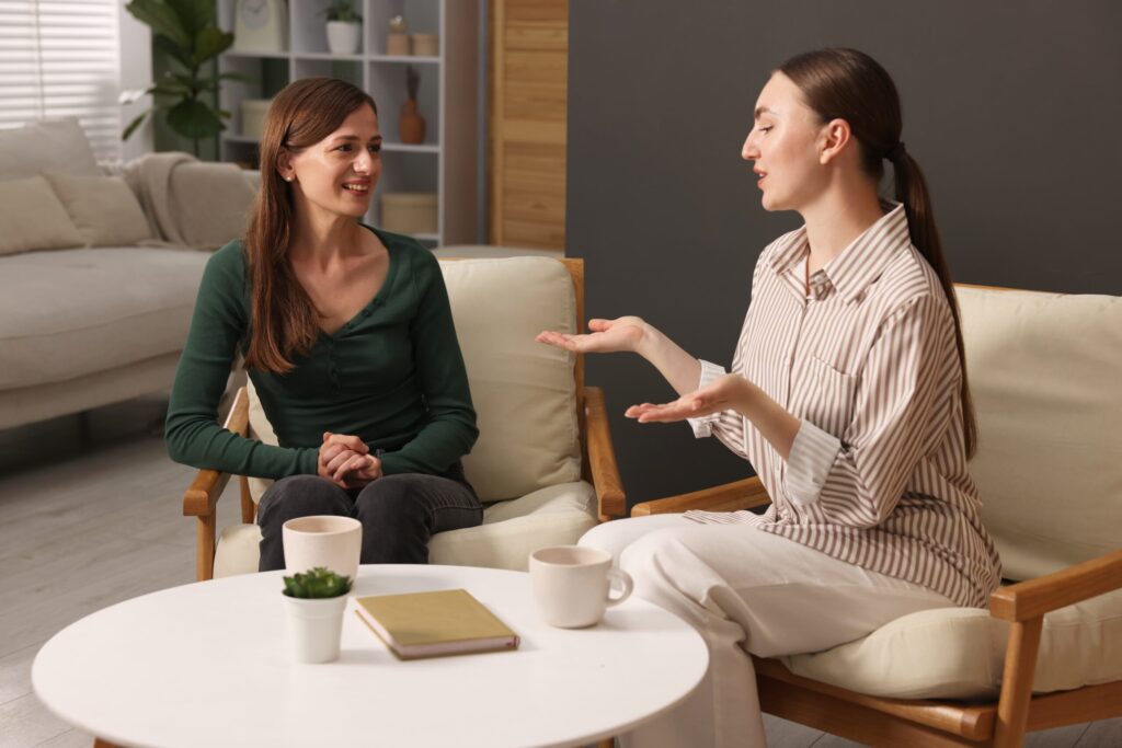 Two people sitting in comfortable chairs in a warm, clean, modern therapy room setting, engaged in a focused, hopeful conversation